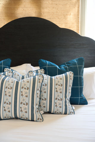 A neatly made bed with a dark curved headboard and four decorative pillows: two teal grid-patterned ones and two Rose Garden Pillows with blue floral designs, all arranged on crisp white bedding.