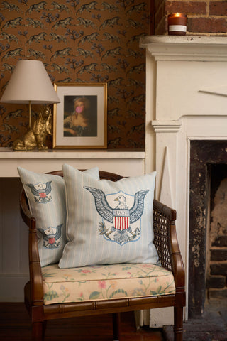 A wooden chair with vintage floral upholstery holds two One Nation Pillows, set beside a white fireplace. On a nearby desk are a lamp, gold dog figurine, framed portrait, and a lit candle.