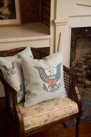 A wooden chair with a vintage floral pillow and two light blue One Nation Pillows featuring an eagle and a shield labeled “USA.” A fireplace and framed picture appear in the background.