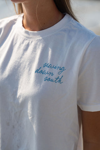 A person wearing the Women’s Dock Days Tee, a white T-shirt featuring blue “Sewing Down South” text from the Shore Capsule Collection. The image is cropped to show only the upper torso, partially illuminated by sunlight.