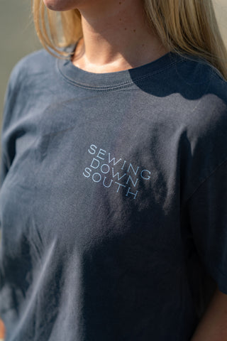 A person is seen wearing the Dock Days Tee in dark gray, featuring SEWING DOWN SOUTH printed in white on the chest. Part of the shore capsule collection, this tee offers an oversized fit and is shown on a model with only the upper torso and part of their face visible.
