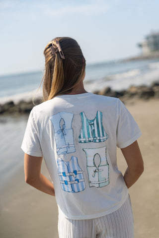 A woman with a hair clip stands on the beach, facing away. She wears the Women’s Dock Days Tee—a white T-shirt with four illustrated blue and white patterned tank tops on the back—paired with light striped pants. Waves and rocks surround her, conveying effortless style inspired by the shore capsule collection.
