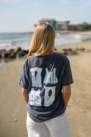 A person with long blonde hair stands on a sandy beach facing the ocean. They are wearing the Dock Days Tee, which features an oversized fit and four cartoon swimsuit designs on the back, from the Shore Capsule Collection, paired with white shorts. The sky is clear and a building can be seen in the distance.