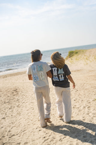 Two people stroll arm in arm along a sandy beach toward the ocean, both dressed in light pants and oversized Dock Days Tees from the Shore Capsule Collection, sunlight illuminating their path on a clear day.