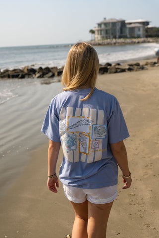 A woman with blonde hair, facing away from the camera, walks along a sandy beach near rocks wearing the Coastal Curio Tee—an oversized, boxy-fit light blue t-shirt made from heavyweight cotton—paired with white shorts. In the background, a house and water are visible.