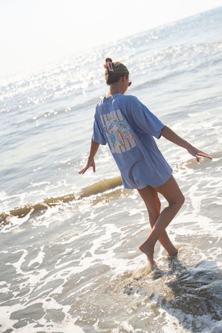 A person with their hair in a bun walks barefoot through shallow ocean water, arms outstretched for balance under bright sunlight, wearing the Coastal Curio Tee—an oversized, boxy-fit blue shirt crafted from heavyweight cotton.