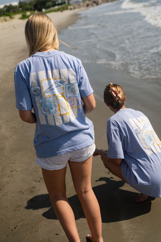 On a sunlit, sandy beach, two women wear light blue Coastal Curio Tees with seashell artwork displayed on the back. Featuring an oversized, boxy fit for a relaxed look, one woman stands facing the ocean while the other crouches to touch the sand near the water’s edge.