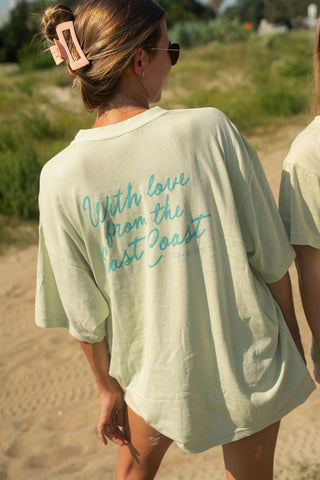 A woman with a pink hair clip stands outdoors on a sandy path surrounded by greenery, wearing the light-colored, oversized “With Love from the East Coast Tee,” featuring blue script on the back. She faces away from the camera, embodying the relaxed spirit of coastal towns.