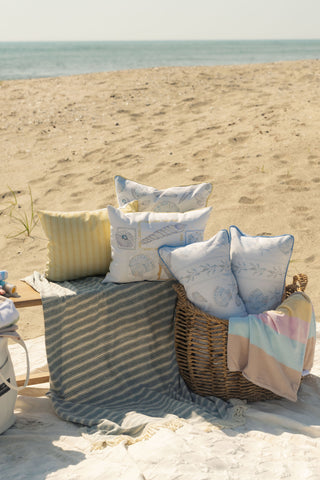 A basket of pillows, featuring the Outdoor Coastal Curio Lumbar Pillow and a pastel blanket, sits on a sandy beach next to a striped rug, with the ocean and sky in the background.