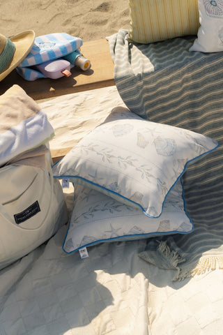 Two Outdoor Sullivan’s Shore Pillows in pale blue with seashell patterns rest on a sandy beach setup, surrounded by a striped blanket, a tote bag, folded towels, and a sun hat under bright sunlight.