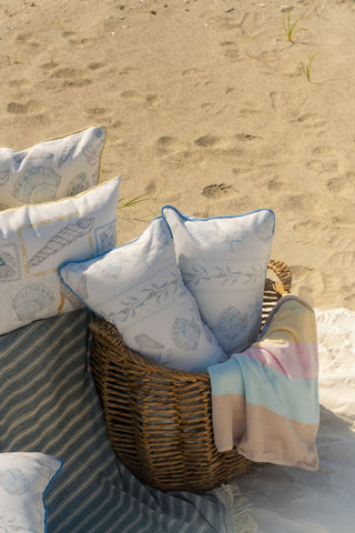 A wicker basket holds patterned pillows and a pastel-striped blanket on a sandy beach, with the Outdoor Sullivan’s Shore Extended Lumbar Pillow and a striped blanket nearby.