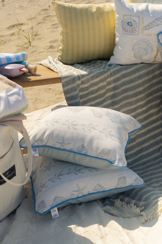 Two white Outdoor Sullivan’s Shore Pillows with blue piping and seashell patterns rest on a blanket at the beach. Nearby are a striped coastal outdoor pillow, a yellow pillow, a beach bag, and a wooden tray holding sunblock and sunglasses.