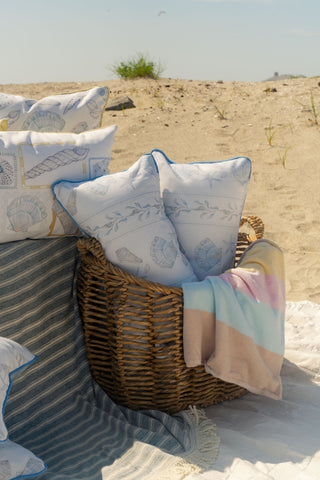 A wicker basket holding the Outdoor Sullivan’s Shore Extended Lumbar Pillow and a pastel-striped blanket sits on a sandy beach, next to striped rugs and more pillows, with dune grass and blue sky in the background.