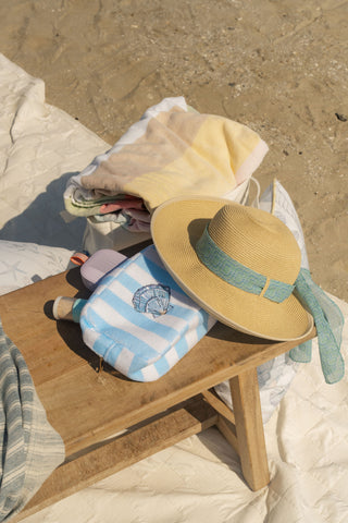 A straw hat, the Weekend Getaway Striped Terry Cosmetic Bag, sunscreen, and folded towels rest on a wooden beach table, with sand and a quilted blanket in the background.