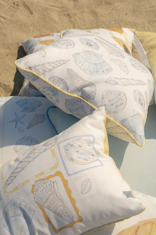 Three Outdoor Washed Ashore Pillows, adorned with delicate seashell illustrations in pastel blue, yellow, and beige tones, are set on sand and a light blanket—washable and perfect for enhancing your coastal outdoor pillow collection.