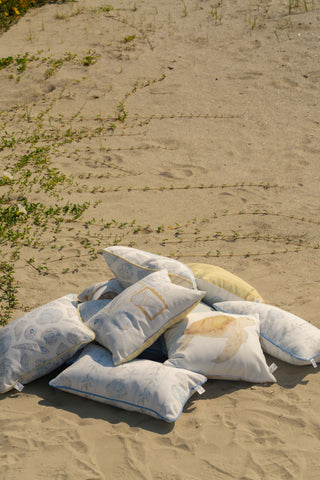 A stack of light-patterned decorative pillows, featuring the Outdoor Tidal Travelers Lumbar Pillow with its charming coastal design, rests on sandy ground amid sparse green plants and faint footprints in the background.