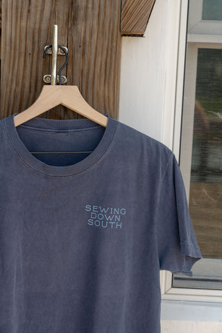 A blue-gray Dock Days Tee featuring the SEWING DOWN SOUTH logo hangs on a wooden hanger in front of a rustic wooden wall and window, highlighting its relaxed, oversized fit as part of the shore capsule collection.