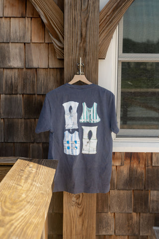 The Dock Days Tee, shown here in gray with four illustrated tank tops on the front, hangs on a wooden hanger outside beside a shingled wall and window, sunlight creating gentle shadows. Designed with an oversized fit, this tee is part of the Shore Capsule Collection.