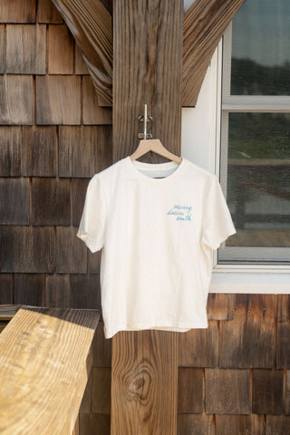 The Women’s Dock Days Tee from the Shore Capsule Collection hangs on a wooden hanger against an outdoor wooden wall near a window, with sunlight casting gentle shadows across the scene.