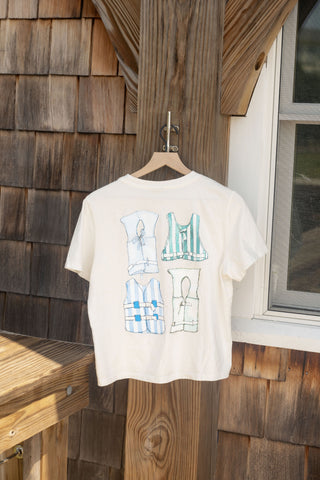 The Women’s Dock Days Tee from our Shore Capsule Collection, featuring four outlined life jacket illustrations, hangs on a wooden hanger outdoors against a shingled wall beside a white-framed window—perfect for those relaxed dock days and boat outings.
