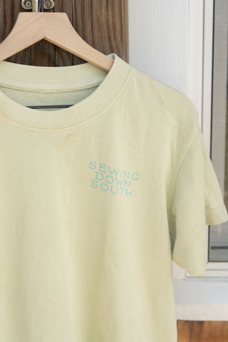 The With Love from the East Coast Tee—a pale yellow shirt from the Shore Capsule Collection featuring “SEWING DOWN SOUTH” printed in light blue—hangs on a wooden hanger against a wooden door, perfectly capturing the relaxed spirit of coastal towns.