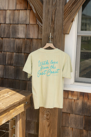 The With Love from the East Coast Tee, in a pale yellow shade, hangs on a wooden hanger outdoors. Blue text reads “With love from the East Coast” across the relaxed fit shirt, a part of our Shore Capsule Collection. Sunlit wood shingles, a window, and wooden railings in the background evoke the breezy charm of coastal towns.