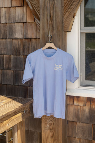 A light blue Coastal Curio Tee, featuring an oversized boxy fit, hangs from a wooden hanger outdoors on a wooden post beside a window, with sunlight creating soft shadows.
