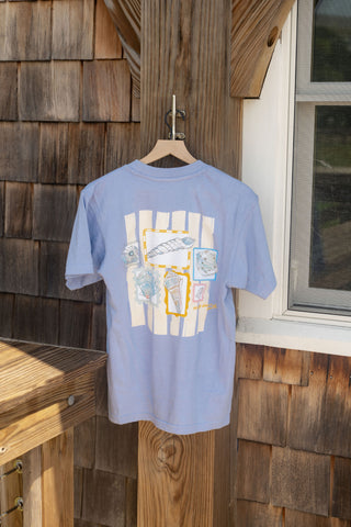 A light blue Coastal Curio Tee is displayed on a wooden hanger outside, featuring fish and seashell artwork, positioned near a window and a wood-paneled wall.