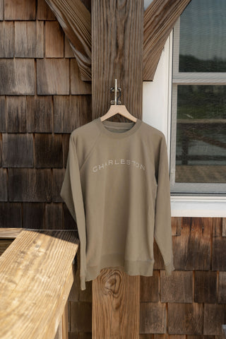 A unisex Charleston Terry Crewneck Sweatshirt in khaki, featuring “CHARLESTON” across the chest, hangs on a wooden hanger by a rustic shingle wall next to a window—an ideal choice for your next go-to coastal sweatshirt.