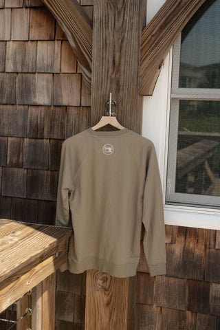 A tan Charleston Terry Crewneck Sweatshirt is displayed on a wooden hanger against a wood-paneled outdoor wall beside a white-framed window. The unisex relaxed fit Charleston Terry Crewneck Sweatshirt brings effortless style to any setting.