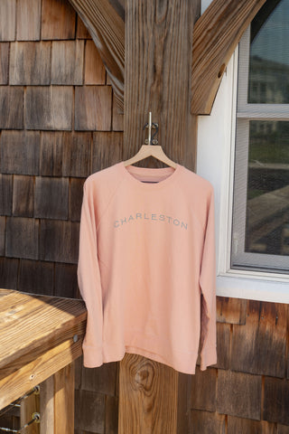 A Charleston Terry Crewneck Sweatshirt in light pink, featuring “CHARLESTON” printed across the chest, is displayed on a wooden hanger outside against a weathered wooden wall near a window. This unisex, relaxed-fit sweatshirt blends comfort with coastal style, making it ideal for any laid-back occasion.