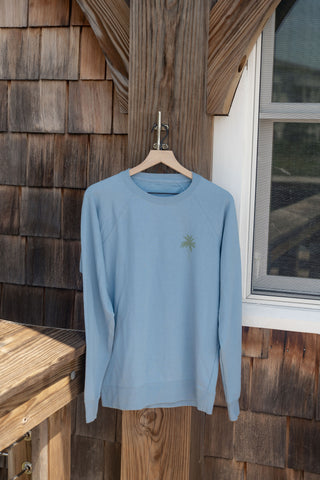 The Weekends on the Shore Terry Crewneck Sweatshirt, in light blue terry fabric and featuring a small green palm tree graphic, hangs on a wooden hanger outside against a wooden wall near a window. This unisex relaxed fit is perfect for weekends spent by the shore.
