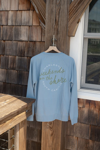 The Weekends on the Shore Terry Crewneck Sweatshirt, in a light blue terry fabric, hangs on a wooden hanger outdoors. Featuring “Charleston, Weekends on the Shore, South Carolina” printed across the back in white and green lettering, this unisex relaxed fit is perfect for breezy coastal days.