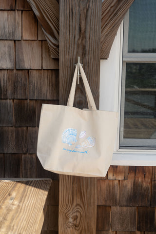 A beige canvas Coastal Curio Tote, featuring delicate seashell artwork, hangs on a wooden post beside a window on a shingled building. This tote brings breezy seaside charm to any setting.