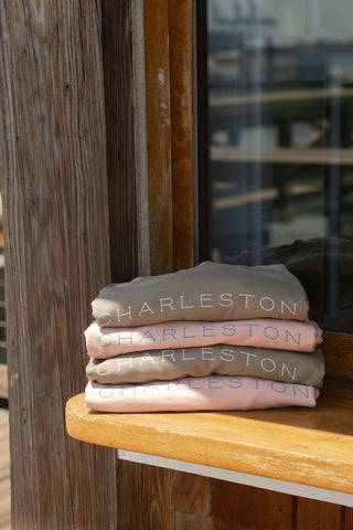 Three Charleston Terry Crewneck Sweatshirts in neutral colors, each featuring “CHARLESTON” printed across the front, are stacked on a wooden ledge by a window with rustic wooden paneling.