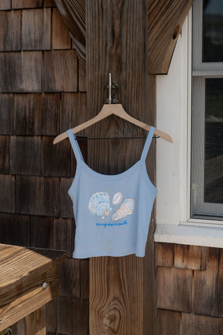 The Coastal Curio Cropped Tank, featuring seashell artwork and ocean youth text on a light blue cropped design, hangs on a wooden hanger outside on a shingled wall near a window, making it the perfect piece of casual beachwear.