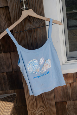 The Coastal Curio Cropped Tank—a light blue cropped top featuring seashell artwork and “sewing down earth” text—hangs on a wooden hanger by a window, with wooden siding in the background, making it perfect for casual beachwear.