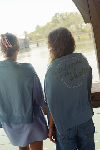 Two people stand side by side facing a waterfront view. Both wear light blue shirts; one, the Weekends on the Shore Terry Crewneck Sweatshirt that reads “Weekends on the Shore, Charleston, South Carolina.” Their hair is loose, and one has a large hair clip.