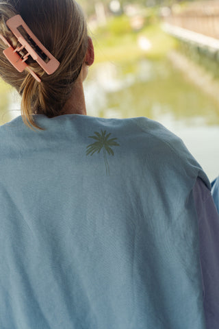 A person with light brown hair in a pink hair clip is standing near water with greenery in the background, wearing the Weekends on the Shore Terry Crewneck Sweatshirt—a light blue unisex sweatshirt featuring a small palm tree graphic on the back—perfect for relaxed weekends by the shore.