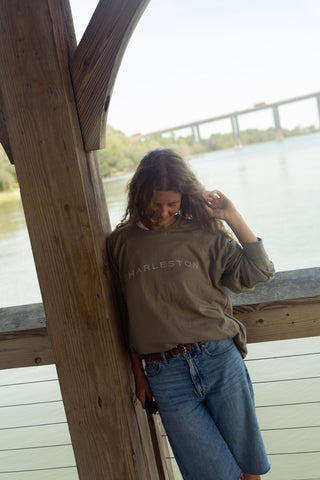 Wearing the Charleston Terry Crewneck Sweatshirt and denim shorts, a woman leans against a wooden railing by the water, smiling as she looks down, with a bridge in the background.