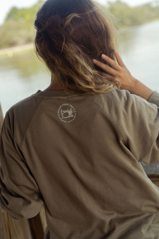 A person with wavy brown hair faces away while sitting by the water, touching their head. They are wearing a tan Charleston Terry Crewneck Sweatshirt featuring a circular logo on the back. Trees and blurred greenery can be seen in the background.