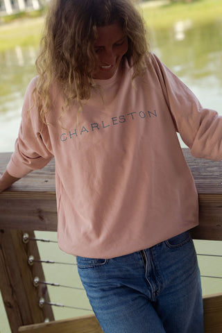 A person with wavy hair wearing the Charleston Terry Crewneck Sweatshirt in pink and blue jeans leans against a wooden railing, smiling and looking down, with water and greenery in the background.