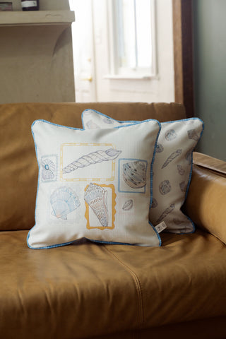 Two decorative pillows with seashell illustrations rest on a tan leather couch. The front pillow, the Coastal Curio Pillow, features blue trim and showcases various seashell drawings within colored frames; the back pillow displays a simpler seashell pattern. Both coastal pillow covers make perfect accents for seaside-inspired decor.
