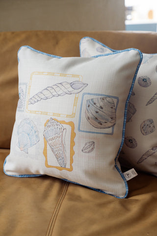 A square Coastal Curio Pillow with blue piping and illustrated seashell designs sits on a tan couch, while another matching pillow is partially visible in the background.