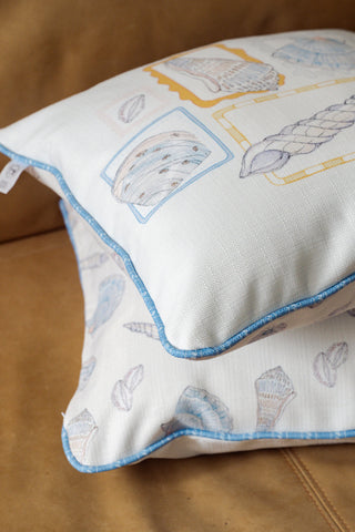 Two Coastal Curio Pillows rest on a tan leather surface, each featuring pastel-colored, ocean-themed patterns for a subtle yet charming coastal look. The pillows are stacked neatly, showcasing their white fabric adorned with blue trim and seashell designs.