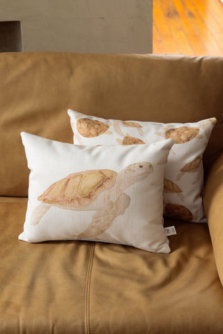 Two Tidal Travelers Prefilled Lumbar Pillows featuring charming brown sea turtle illustrations rest on a tan leather couch. The white pillows are set against a beige wall and wooden floor, with sunlight streaming in to create a serene, coastal vignette.