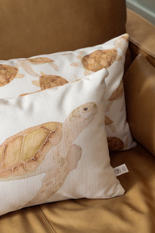 Two cream-colored pillows with brown sea turtle illustrations rest on a light brown leather armchair. One Tidal Travelers Prefilled Lumbar Pillow is in the foreground, showing a turtle in detail, while the other coastal throw pillow is angled behind it.