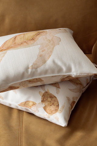 Two Tidal Travelers Prefilled Lumbar Pillows with light brown sea turtle patterns rest on a tan leather couch, creating the perfect coastal accent pillow arrangement.