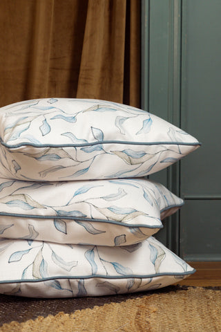 Three Shoreline Vines Pillows, featuring a white base with a blue and gray abstract leaf pattern reminiscent of coastal décor, are stacked on a woven rug in front of a brown curtain and a muted blue-green wall and door.