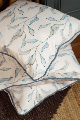 Two decorative Shoreline Vines Pillows, each featuring a blue and gray leaf pattern, piped edges, and a soft, artistic design on a subtle coastal cover, rest on a textured brown rug.